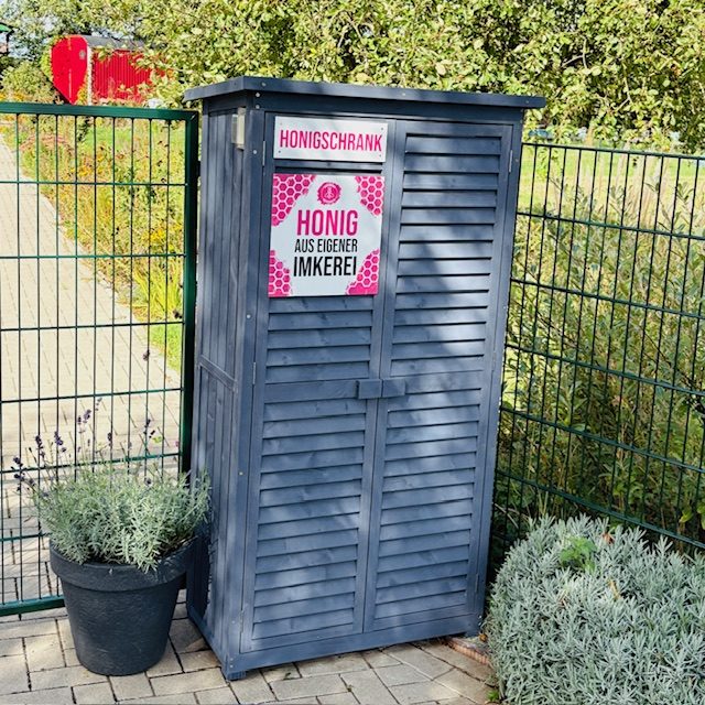 Grauer Schrank mit beschrifteter Tafel, umgeben von Pflanzen und einem Zaun.