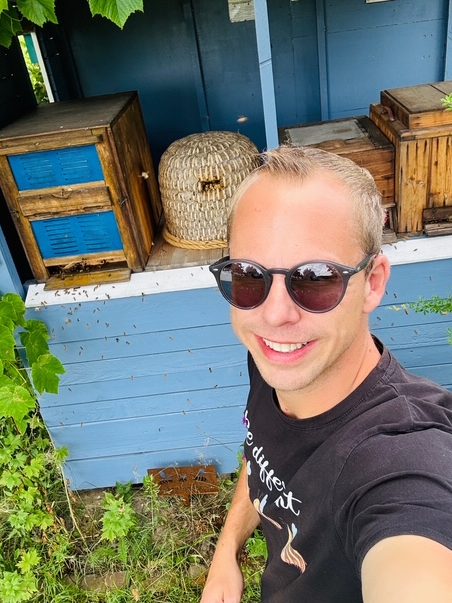 Timo Weitz Bio-Imkerei Bienenkönig Niederrhein Timo Weitz mit Sonnenbrille steht vor einem bunten Bienenstock und grünen Pflanzen.
